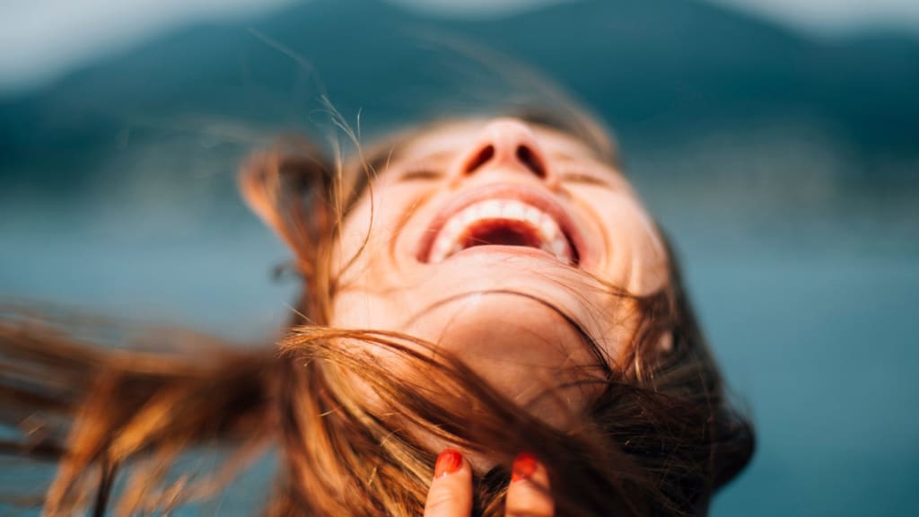 Close-up of a woman’s face, head tilted back and mouth open in laughter, capturing a joyful moment that could follow therapy or counseling, with wind blowing her hair and a blurred background.